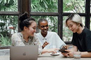 Three young professionals smiling while looking at a phone and laptop, representing effortless money management and financial automation.