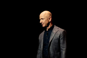 A man standing on a dark stage wearing a blazer, speaking with a spotlight on him.