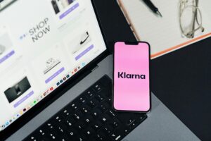 Pink smartphone showing the Klarna logo on a desk beside a laptop displaying an online store checkout page.