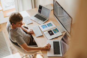 Man working from home at a desk surrounded by multiple screens, papers, and a laptop.